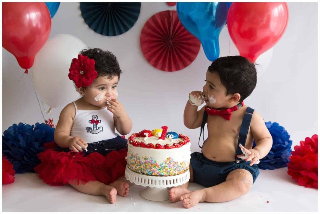 Twin brother and sister sharing one first birthday cake in front of red and blue balloons