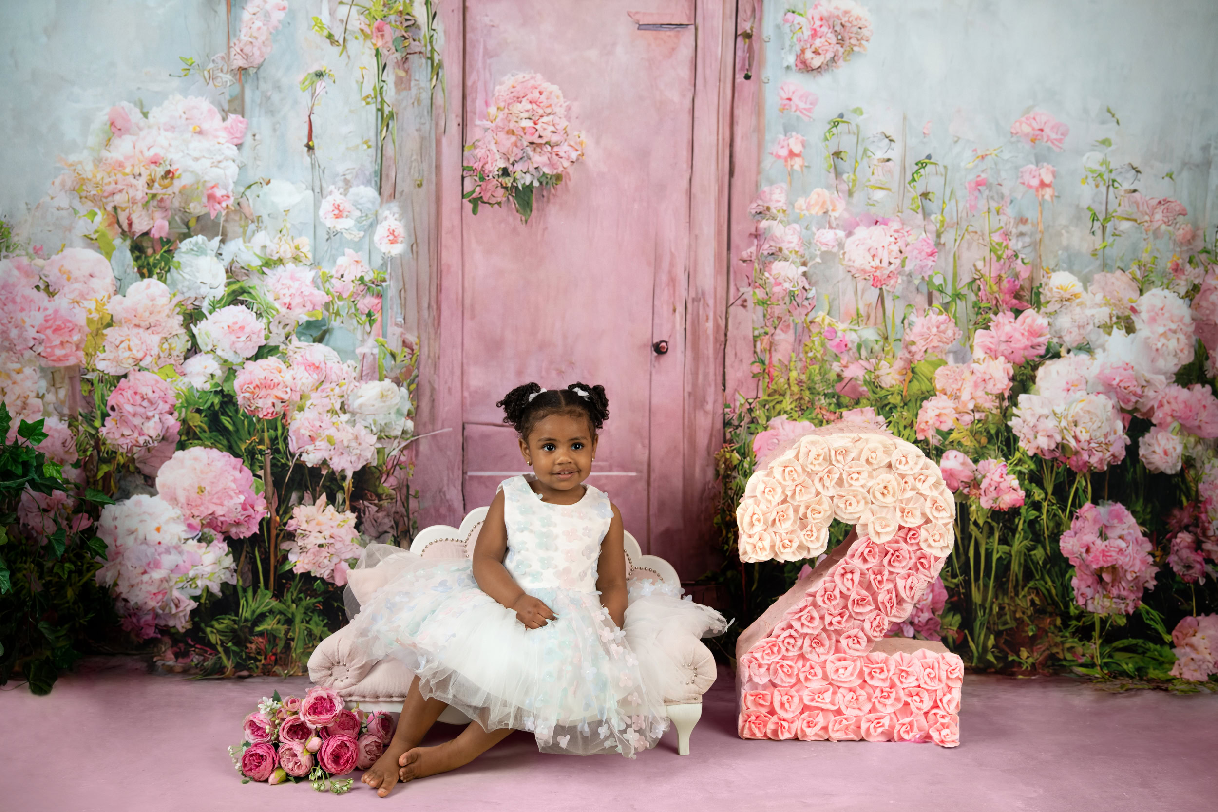A little girl in a dress sitting in front of a pink door with a number two.
