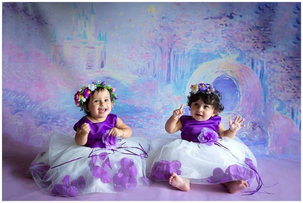 Twin sisters in purple and white tutu dresses with flower crowns in front of a fairytale castle backdrop