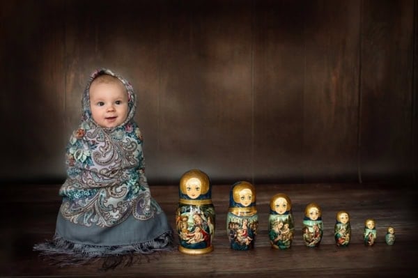 Baby wrapped in a colorful traditional Russian headscarf, sitting on a dark wooden floor next to a row of ornate Russian nesting dolls, photographed in a creative newborn portrait session in Washington DC.