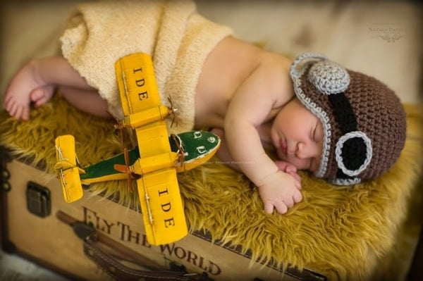 Newborn baby boy sleeping on his side on a soft yellow faux fur blanket placed over a vintage travel suitcase with &ldquo;Fly the World&rdquo; text, wearing a handmade brown and gray crochet aviator hat with goggles, next to a miniature yellow and green biplane toy, photographed during a themed newborn photography session in Washington, DC.
