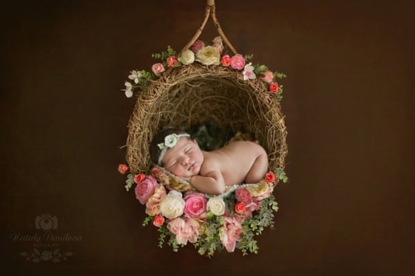 Newborn baby girl sleeping on her side inside a round hanging nest made of twigs and decorated with pink, coral, and white flowers, wearing a delicate floral headband, photographed against a warm brown background during a professional newborn session in Washington, DC.