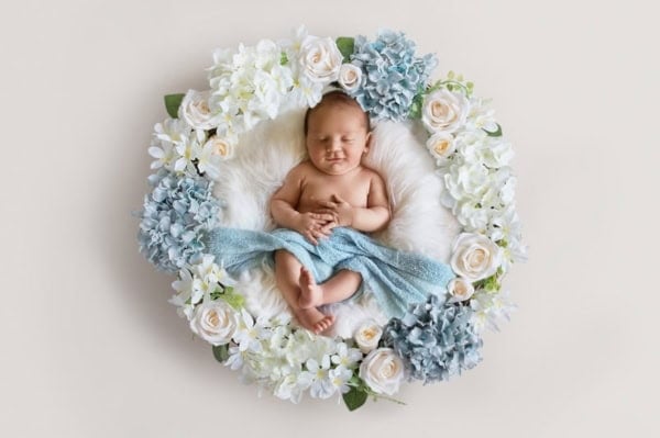 Top-view newborn portrait of a sleeping baby lying on a fluffy white blanket, loosely draped in a light blue wrap, with hands gently clasped over the chest. The baby is surrounded by a perfectly arranged floral wreath featuring white roses, daisies, and clusters of blue hydrangeas, creating a soft and dreamy composition against a light beige background.