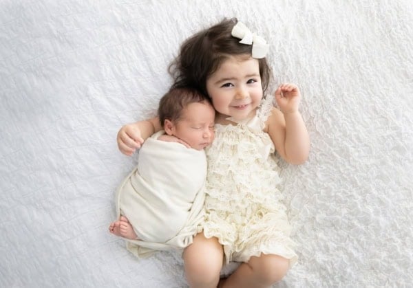 Big sister hugging and smiling next to her swaddled newborn baby brother on a white blanket, captured by Washington DC newborn photographer.