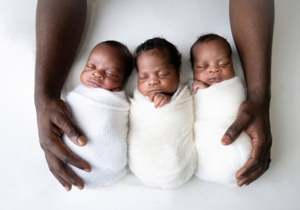 Three African American newborn triplets, each swaddled in soft white wraps, sleeping side by side on a white background, with their father&rsquo;s hands cradling them from both sides, captured during a professional newborn photography session in Washington, DC.