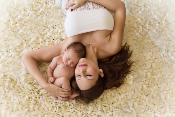 Washington DC newborn photographer top-view portrait of a mother in a white strapless dress lying on a textured bed of cream-colored petals, holding her naked newborn baby against her chest. Both mother and baby have their eyes closed, sharing a peaceful and intimate bonding moment.