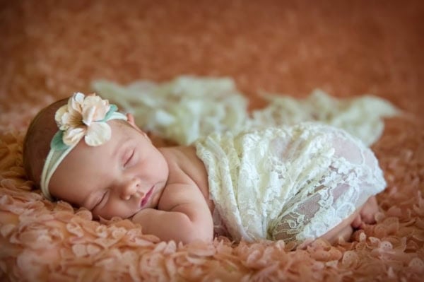 Newborn baby girl in white lace outfit with flower headband sleeping on peach floral blanket &mdash; Northern Virginia newborn photography.