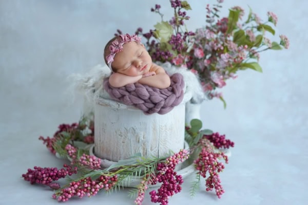 Newborn baby girl sleeping in a whitewashed rustic bucket, wrapped in a chunky purple knit blanket with a matching floral headband, surrounded by lush purple berries, pink blossoms, and greenery &mdash;