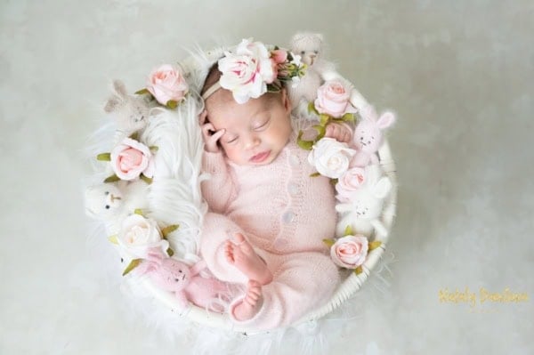 Washington DC newborn photographer top-view image of a newborn baby girl wearing a soft pink knitted romper and a floral headband, sleeping peacefully in a white round basket lined with fluffy faux fur. The basket is decorated with light pink roses and small stuffed animals, creating a dreamy, romantic newborn portrait.