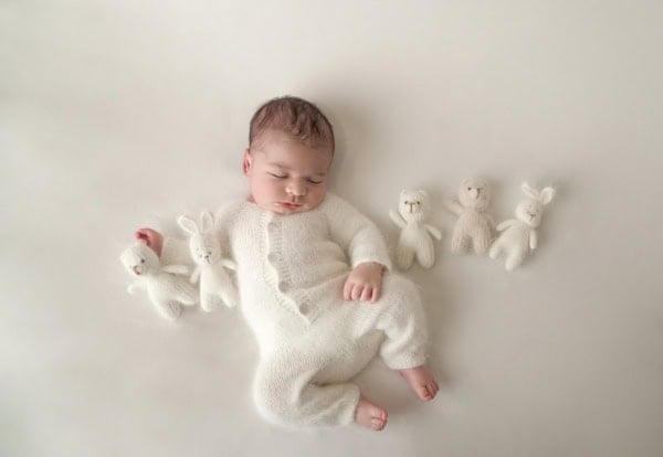 Washington DC newborn photographer studio portrait of a sleeping baby dressed in a cozy white knitted onesie, lying on a light cream background with five small handmade plush toys &mdash; three bunnies and two bears &mdash; neatly arranged around them, creating a serene and heartwarming newborn photo.