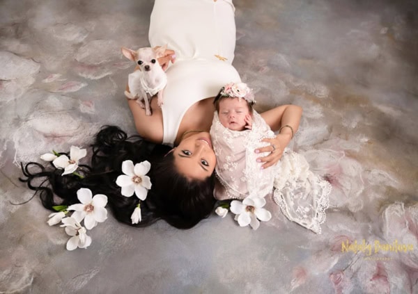 Elegant portrait of a mother in a white dress lying on a soft, hand-painted floral backdrop, holding her newborn wrapped in a delicate lace swaddle, with magnolia flowers in her hair and a small Chihuahua sitting next to her &mdash; captured in a luxury family photography studio in Vienna, Virginia.