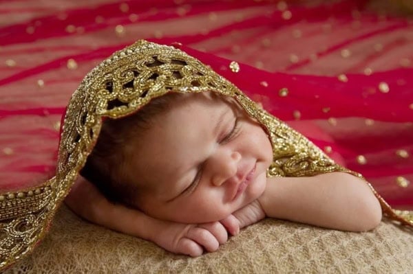 Newborn baby wrapped in traditional gold and red fabric, sleeping on a soft beige surface &mdash; Washington DC newborn photographer.