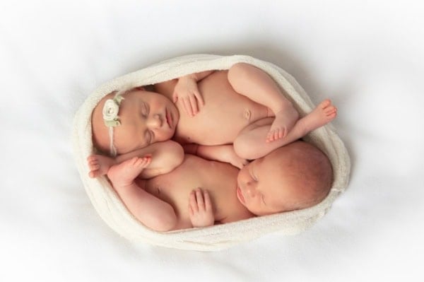 Washington DC newborn photographer top-view photograph of two sleeping newborn twins wrapped together in a creamy white swaddle on a soft white background. The baby girl, wearing a small white flower headband, rests with her head tilted toward her twin brother, who lies beside her with relaxed hands and feet. Their tiny limbs are gently intertwined, creating a heartwarming and intimate pose symbolizing their special bond.