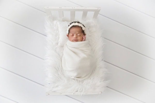 Washington DC newborn photographer top-view image of a newborn baby girl wrapped in a soft white swaddle, lying in a small white wooden bed with a fluffy faux fur blanket. She wears a delicate lace headband and rests her cheek on her tiny hand, creating a peaceful and pure newborn portrait.