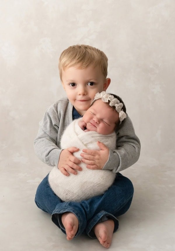 Older brother sitting barefoot on the floor holding his swaddled newborn sister wearing a floral headband &mdash; Washington DC newborn photographer.