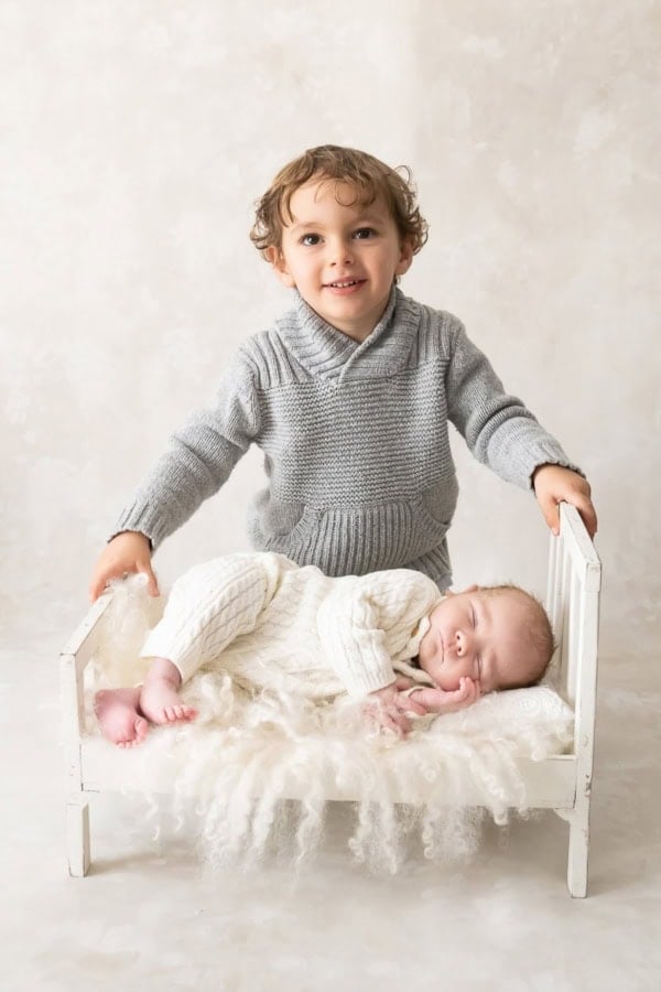 Smiling toddler in gray sweater standing beside newborn sleeping on a small white bed &mdash; Washington DC newborn photography.