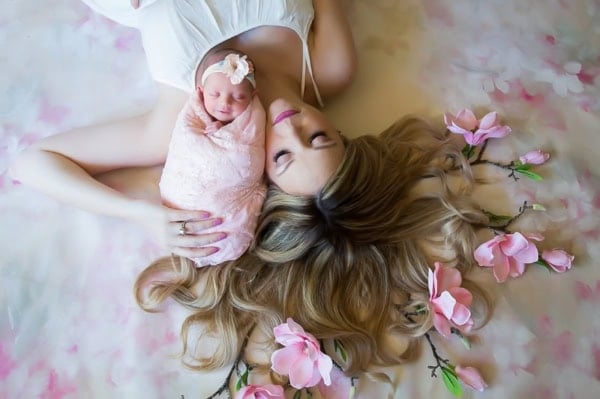 Artistic newborn and mother portrait featuring a blonde-haired mother in a white dress lying on her back with eyes closed, holding her swaddled newborn baby girl dressed in a soft pink wrap and matching floral headband. The scene is styled with the mother&rsquo;s long hair spread out around her, interwoven with pink cherry blossom flowers, creating a dreamy and tender composition.
