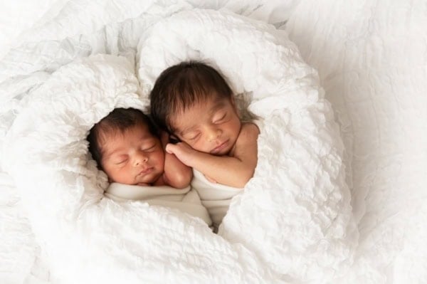 Washington DC twin newborn photographer studio portrait of two sleeping newborns swaddled in soft white wraps, nestled together inside a heart-shaped fold of a textured white blanket, creating a tender and artistic photograph celebrating the love and connection between twins.